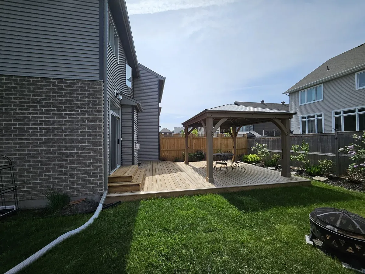 Backyard gazebo sheltering a table and chairs on a deck, viewed across the lawn from the side of the house