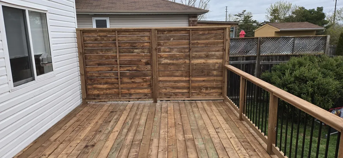 Dark-stained horizontal privacy screen enclosing one end of a back deck beside a white-sided house