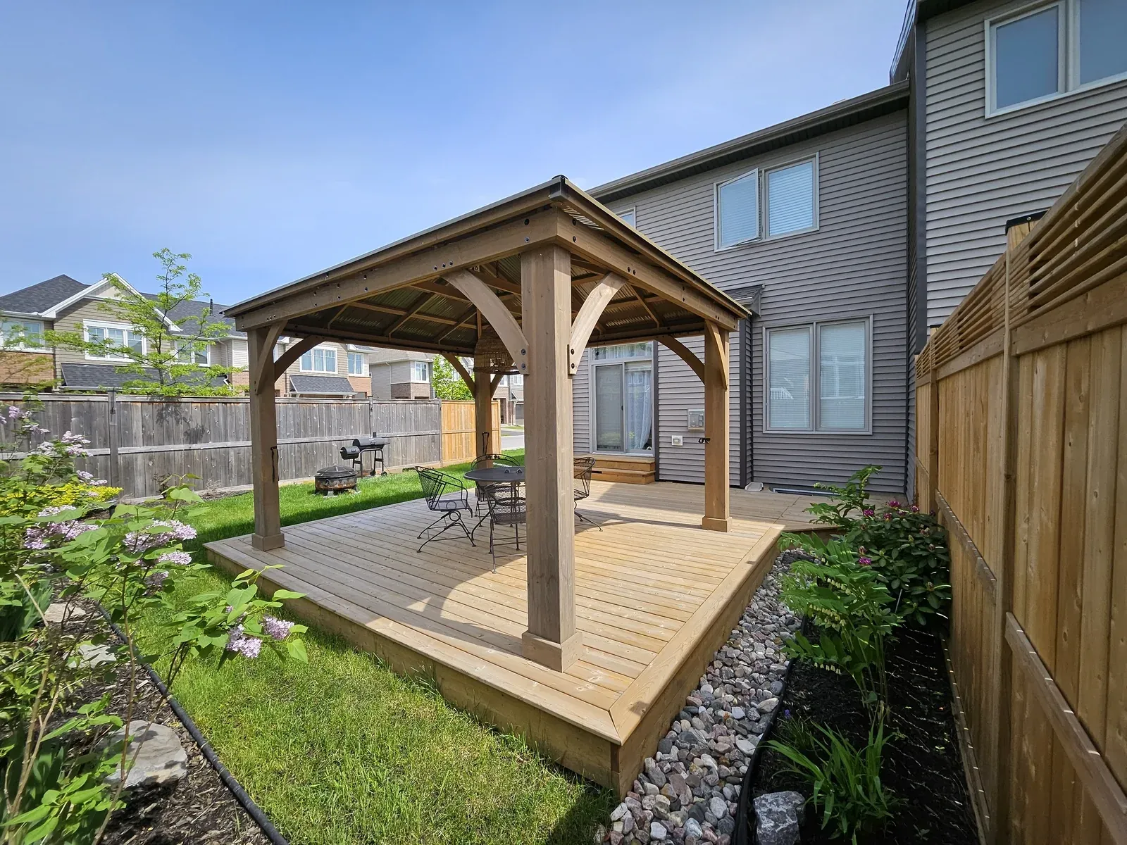 Cedar gazebo with open sides and a steel hip roof standing on a raised cedar deck pad beside a grey-sided house in Newmarket