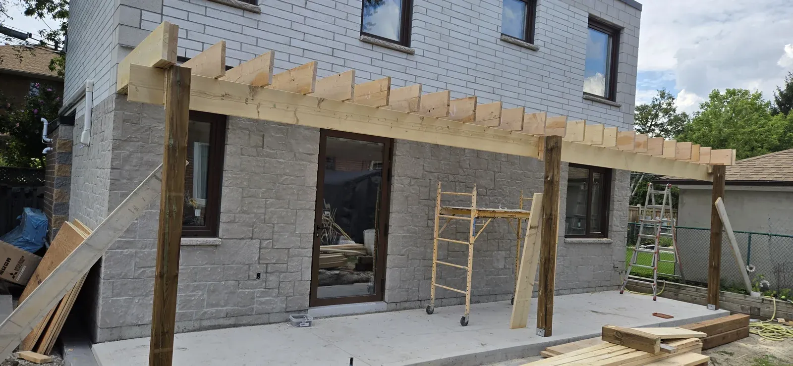 Cedar pergola under construction over a back patio in Newmarket, Ontario, with rafters set on posts against the brick wall of the house